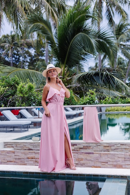 Retrato de uma mulher caucasiana em um vestido longo rosa elegante romântico de férias em um hotel luxuoso e rico, com uma vista incrível de palmeiras tropicais. Mulher com chapéu branco clássico