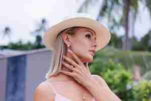 Foto grátis retrato de uma mulher caucasiana em um vestido longo rosa elegante romântico de férias em um hotel luxuoso e rico, com uma vista incrível de palmeiras tropicais. mulher com chapéu branco clássico