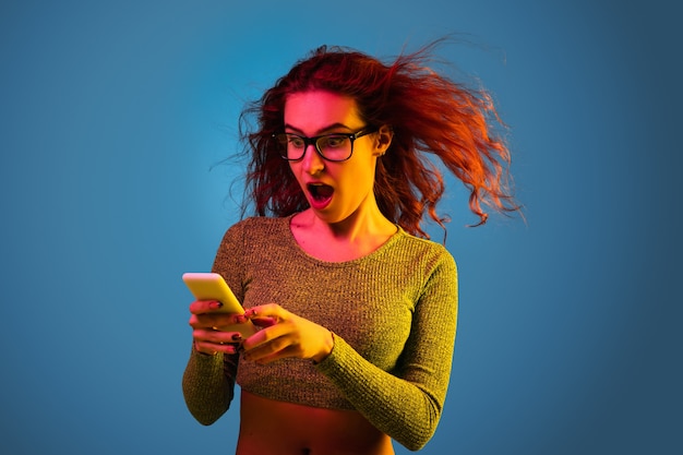 Foto grátis retrato de uma mulher caucasiana isolado em um fundo azul do estúdio em luz de néon. linda modelo feminino com cabelo ruivo. conceito de emoções humanas, expressão facial, vendas, anúncio, aposta. usando smartphone.