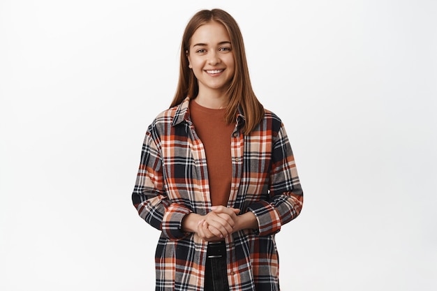 Foto grátis retrato de uma mulher caucasiana simpática e educada, assistente de mulher prestativa, sorrindo agradável, de mãos dadas apertadas sobre o peito, em pé contra uma parede branca