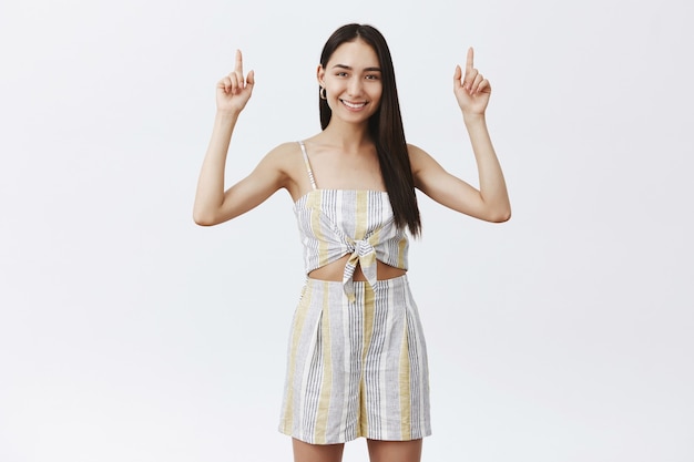 Foto grátis retrato de uma mulher charmosa, simpática e elegante com longos cabelos escuros e verruga sob os lábios, sorrindo amplamente enquanto levanta as mãos e aponta para cima