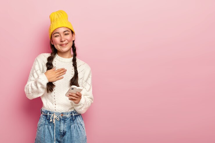 Foto grátis retrato de uma mulher coreana satisfeita com a palma da mão no peito, sendo tocada por palavras comoventes, segura um dispositivo eletrônico moderno, usa um chapéu amarelo, um macacão branco casual e jeans