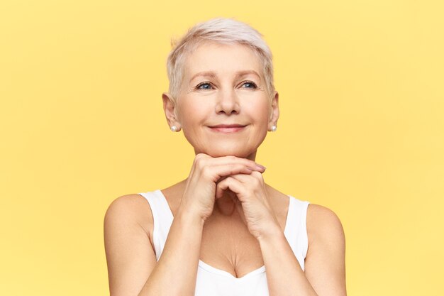 Retrato de uma mulher de meia idade atraente alegre com corte de cabelo curto e elegante e pele bronzeada, colocando as mãos sob o queixo, fazendo anti-envelhecimento rosto lifting massagem.