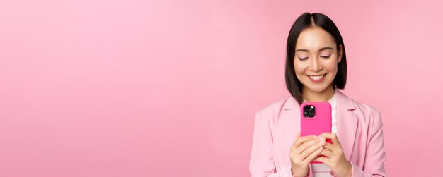 Retrato de uma mulher de negócios sorridente, uma pessoa corporativa asiática usando o aplicativo de smartphone para celular sobre fundo rosa
