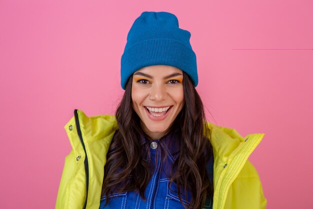 Retrato de uma mulher elegante e sorridente atraente posando com um look de moda de inverno na parede rosa com uma jaqueta amarela brilhante neon, usando um chapéu de malha azul e vestida com roupas quentes