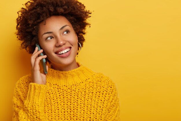 Retrato de uma mulher encaracolada feliz chama um amigo, segura o celular perto da orelha, olha para o lado com um largo sorriso cheio de dentes, usa um macacão amarelo de malha, espaço em branco