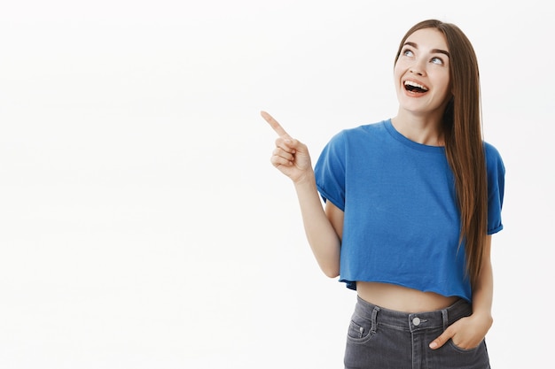 Retrato de uma mulher europeia bonita, divertida e bonita, com uma camiseta azul, apontando e olhando hipnotizada para o canto superior esquerdo com um sorriso encantado