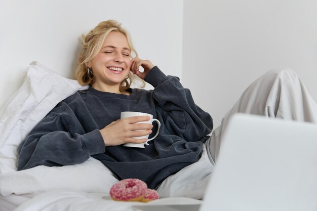 Retrato de uma mulher feliz a desfrutar de um dia de folga em casa, deitada na cama a ver um programa de televisão no portátil.