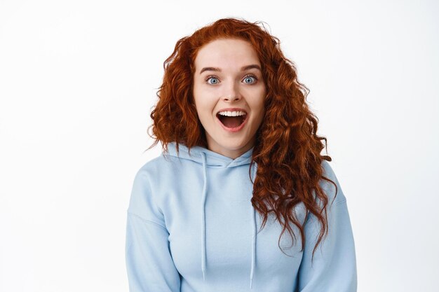 Retrato de uma mulher feliz e animada com cabelo ruivo cacheado, boca aberta, fascinada e maravilhada, olhando fixamente, conferindo notícias incríveis no branco