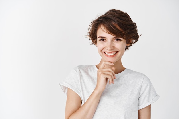 Retrato de uma mulher feliz e sincera sorrindo e tocando o queixo parecendo pensativo em pé contra o fundo branco