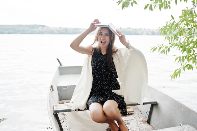 Foto grátis retrato de uma mulher feliz no vestido de bolinhas segurando um livro acima da cabeça enquanto está sentado em um barco velho