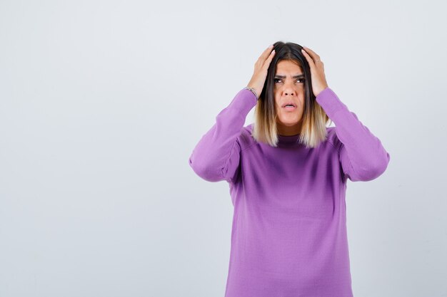 Retrato de uma mulher fofa com as mãos na cabeça em um suéter roxo e olhando para baixo com vista frontal