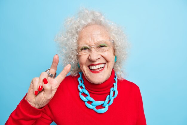 Retrato de uma mulher idosa alegre e bonita sorrindo alegremente fazendo gesto de paz mostra o sinal V vestida com um macacão vermelho e o colar expressa emoções positivas