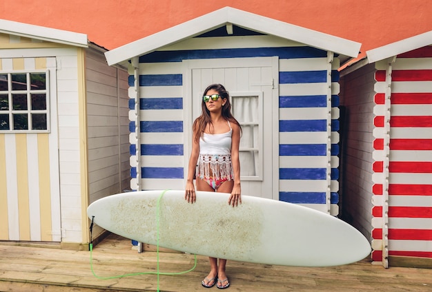 Retrato de uma mulher jovem e bonita surfista com top branco e biquíni segurando a prancha de surf sobre uma praia cabanas listradas. conceito de lazer de verão.