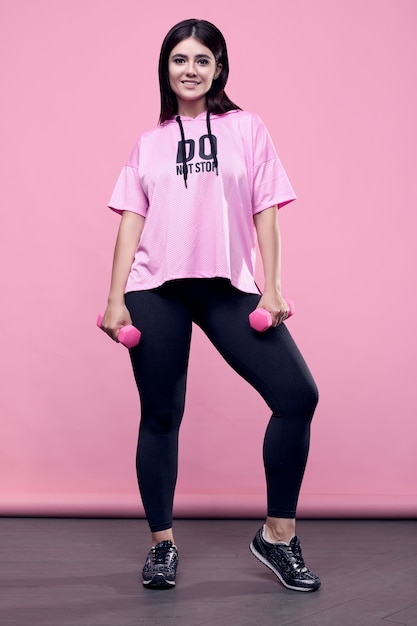 Retrato de uma mulher latina positiva de corpo lindo em um capuz rosa esportes exercitar com halteres em rosa