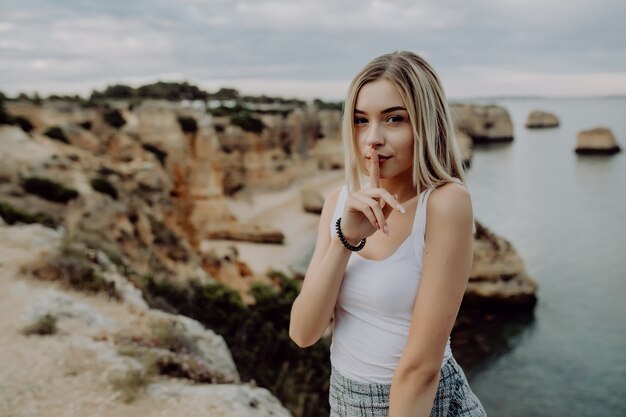 Retrato de uma mulher loira atraente com gesto de silêncio posando na praia rochosa com a paisagem do oceano de beleza.