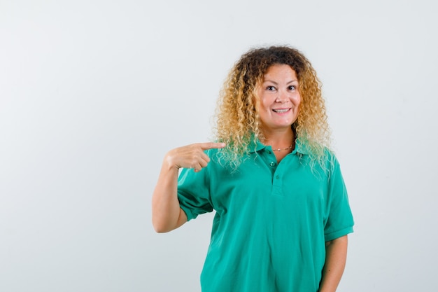 Retrato de uma mulher loira com cabelo encaracolado apontando para si mesma em uma camiseta verde e olhando alegre de frente