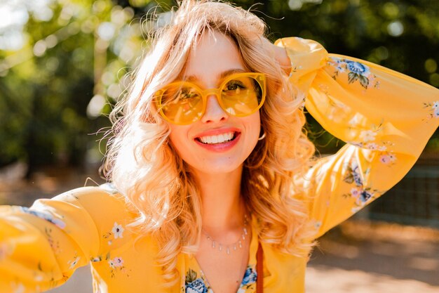 Retrato de uma mulher loira elegante e sorridente bonita em uma blusa amarela usando óculos escuros, fazendo uma foto de selfie