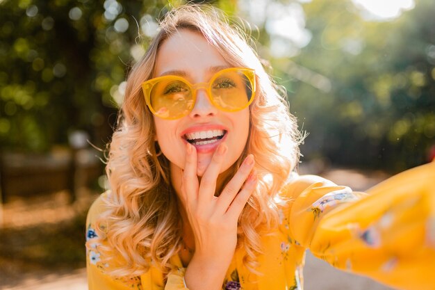 Retrato de uma mulher loira elegante e sorridente bonita em uma blusa amarela usando óculos escuros, fazendo uma foto de selfie