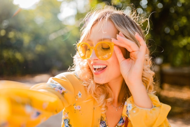 Retrato de uma mulher loira elegante e sorridente bonita em uma blusa amarela usando óculos escuros, fazendo uma foto de selfie