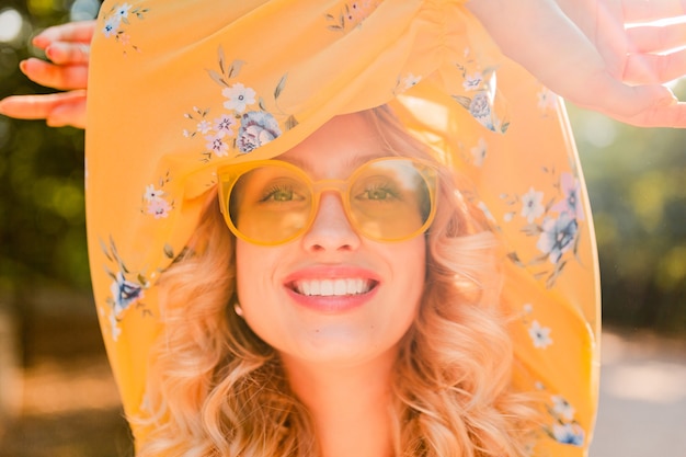 Retrato de uma mulher loira elegante e sorridente com blusa amarela e óculos escuros.