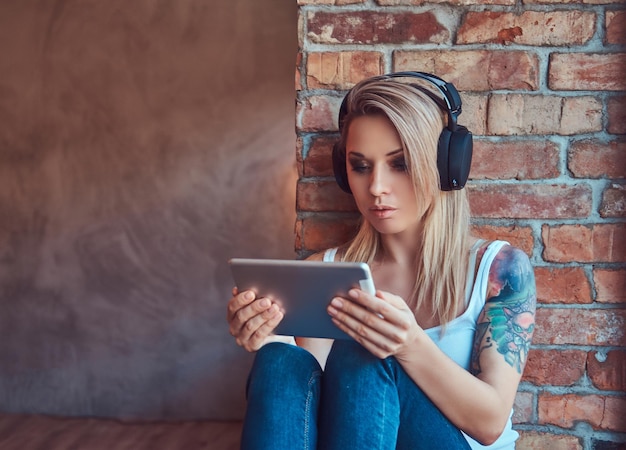 Foto grátis retrato de uma mulher loira tatuada hipster procurando algo em um tablet digital e ouvindo música enquanto está sentado no peitoril da janela em uma sala com um interior loft.