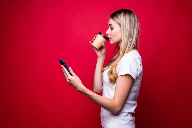 Retrato de uma mulher loira usando o telefone e segurando um café para viagem na parede vermelha