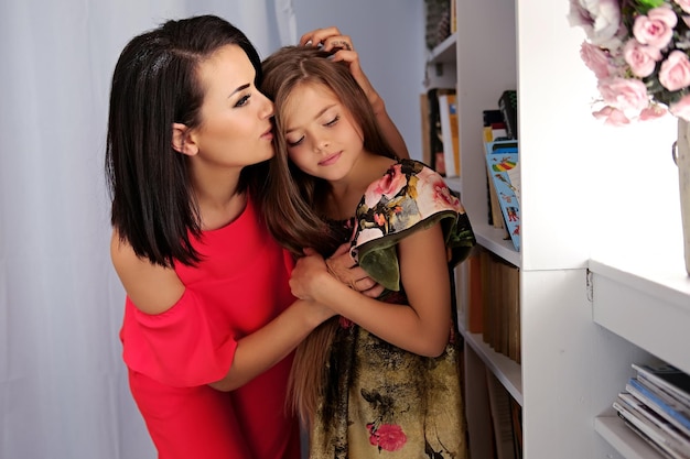 Foto grátis retrato de uma mulher morena atraente vestida com um vestido vermelho, abraçando uma adolescente loira em um quarto.