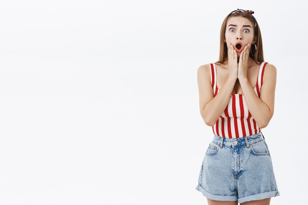 Retrato de uma mulher preocupada, chocada e preocupada, preocupada e preocupada com uma blusa listrada e shorts caindo de queixo tocando o rosto com as mãos saltando os olhos de surpresa