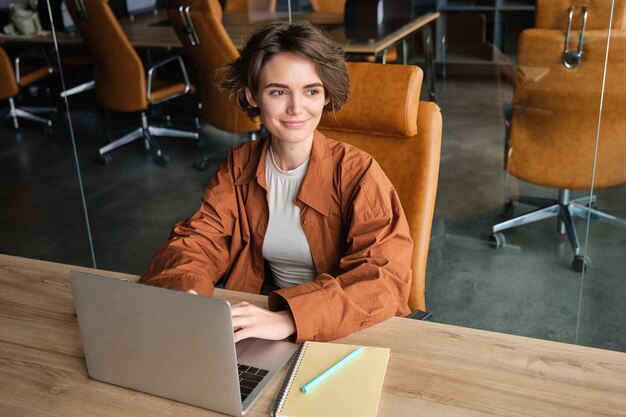 Foto grátis retrato de uma mulher que trabalha no escritório, sentada à mesa com uma programadora de laptop codificando em coworking