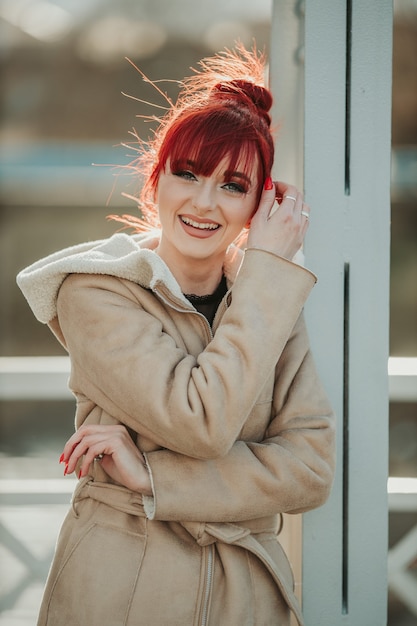 Retrato de uma mulher ruiva com franja apoiada em um pilar de metal vestindo um casaco de inverno quente