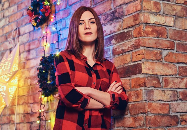 Retrato de uma mulher ruiva vestida com uma camisa de lã vermelha sobre uma parede de decorador de Natal de um tijolo vermelho.