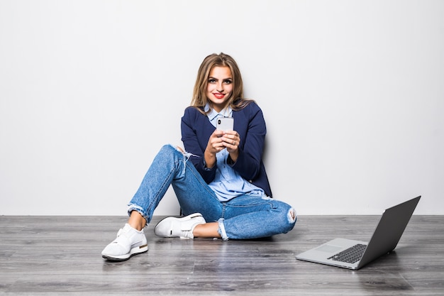 Retrato de uma mulher sorridente, digitando uma mensagem de texto ou rolando na internet usando um telefone celular, sentada sobre uma parede cinza