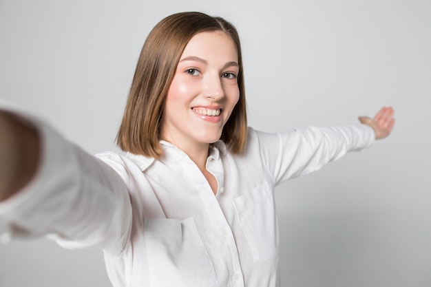 Retrato de uma mulher sorridente e atraente tirando uma selfie enquanto isolado sobre uma parede branca
