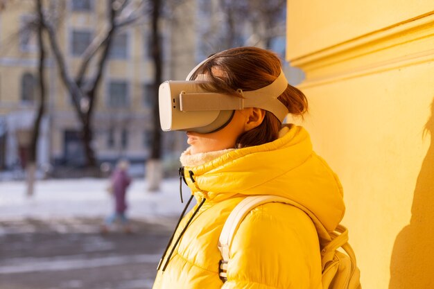 Retrato de uma mulher usando óculos de realidade virtual na rua contra uma parede amarela sob a luz do sol no inverno em roupas quentes