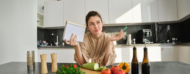 Foto grátis retrato de uma mulher verificando a lista de compras olhando vegetais segurando uma receita de leitura de caderno enquanto