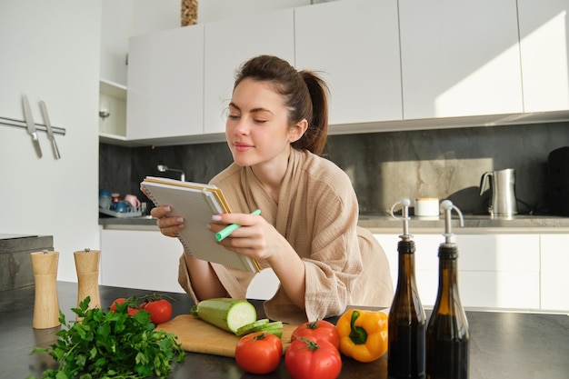 Retrato de uma mulher verificando notas de receita em um caderno em pé na cozinha com legumes cozinhando alimentos