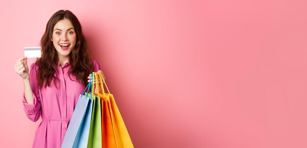 Retrato de uma mulher viciada em compras feliz segurando sacolas de compras e mostrando o cartão de crédito plástico sorrindo espantado em pé contra o fundo rosa