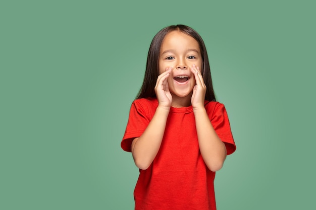 Retrato de uma pequena menina bonita de lado e chamando alguém segurando sua mão perto de sua boca vestindo uma camiseta vermelha, fundo verde
