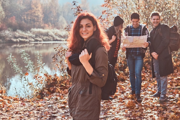 Retrato de uma ruiva encantadora com uma mochila em primeiro plano e um grupo de amigos olhando para o mapa e planejando caminhada em segundo plano na floresta de outono.
