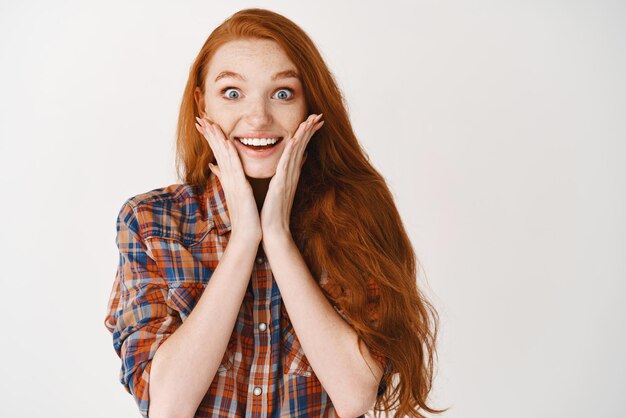 Retrato de uma ruiva feliz ouve notícias incríveis parecendo surpresas e felizes tocando as bochechas e sorrindo para o fundo branco da câmera