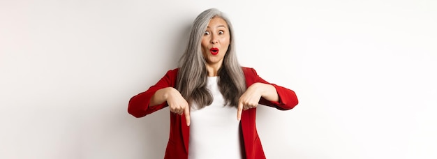 Foto grátis retrato de uma senhora asiática feliz em blazer vermelho mostrando o logotipo apontando os dedos para baixo e sorrindo alegre c