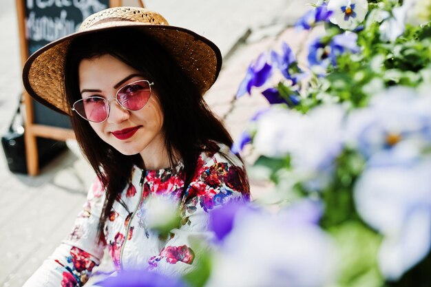 Retrato de verão da menina morena de óculos cor de rosa e chapéu contra loja de flores