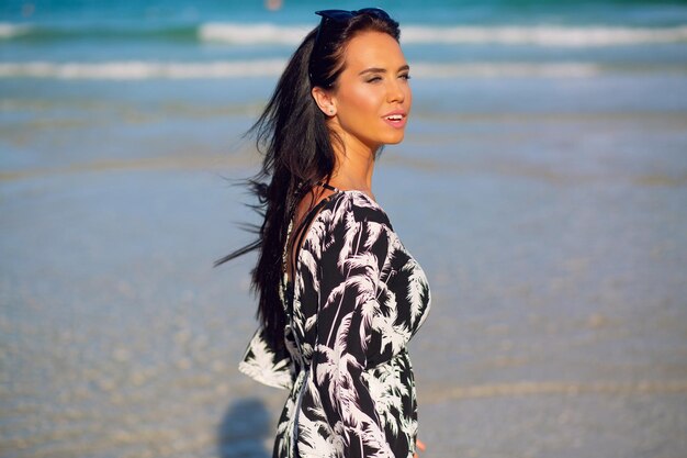 Retrato de verão da mulher bonita morena posando perto do mar azul claro na ilha tropical, vestindo macacão elegante e acessórios. Férias perfeitas perto do oceano.