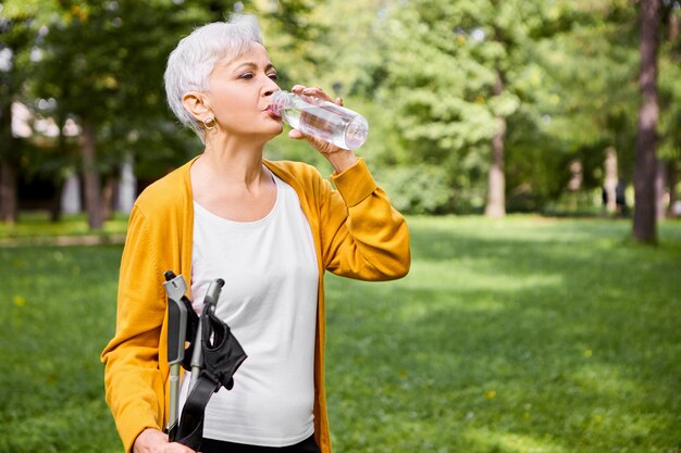 Retrato de verão de uma cansada mulher branca de cabelos grisalhos na casa dos sessenta anos bebendo água de uma garrafa de plástico, se refrescando após a atividade física e posando ao ar livre com bengalas
