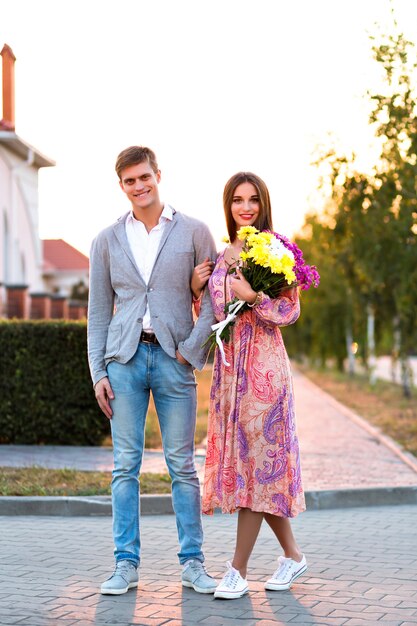 Retrato de verão do incrível casal bonito andando na rua, pôr do sol do campo, roupas elegantes, flores, encontro romântico, amantes bonitos andando na rua.