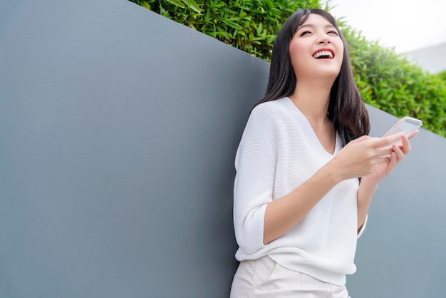 Retrato de vestido casual de cabelo preto comprido asiático atraente, sorriso de mão, uso de smartphone com fundo de paisagem urbana e céu de luz branca, liberdade de copyspace e alegre emocional