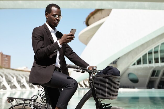Retrato de vista lateral do banqueiro afro-americano moderno ecologicamente consciente, pendulares para trabalhar em bicicleta, tendo um olhar despreocupado e alegre. Atraente empresário preto em roupa formal, andar de bicicleta