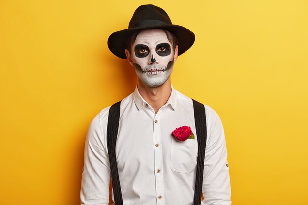 Retrato de zumbi masculino sério usa máscara de caveira, maquiagem horrível, comemora feriado mexicano, usa chapéu preto e camisa branca com suspensórios, tem uma rosa vermelha no bolso, isolada sobre fundo amarelo.