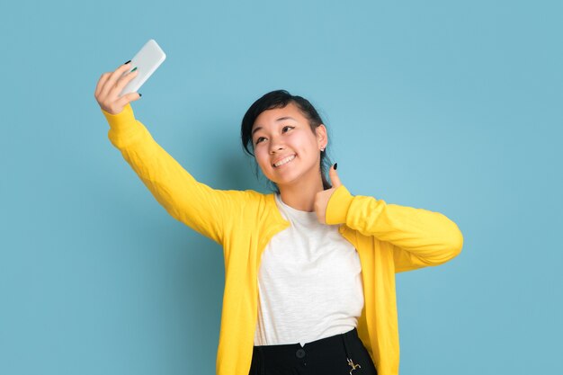Retrato do adolescente asiático isolado no fundo azul do estúdio. Bela modelo moreno feminino com cabelo comprido. Conceito de emoções humanas, expressão facial, vendas, anúncio. Fazendo selfie ou vlog.